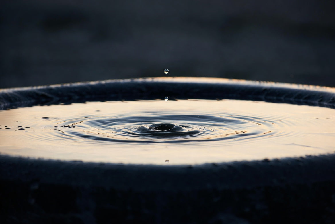 A droplet mid-fall above a dark stone basin of still water, one concentric ripple catching warm dusk light