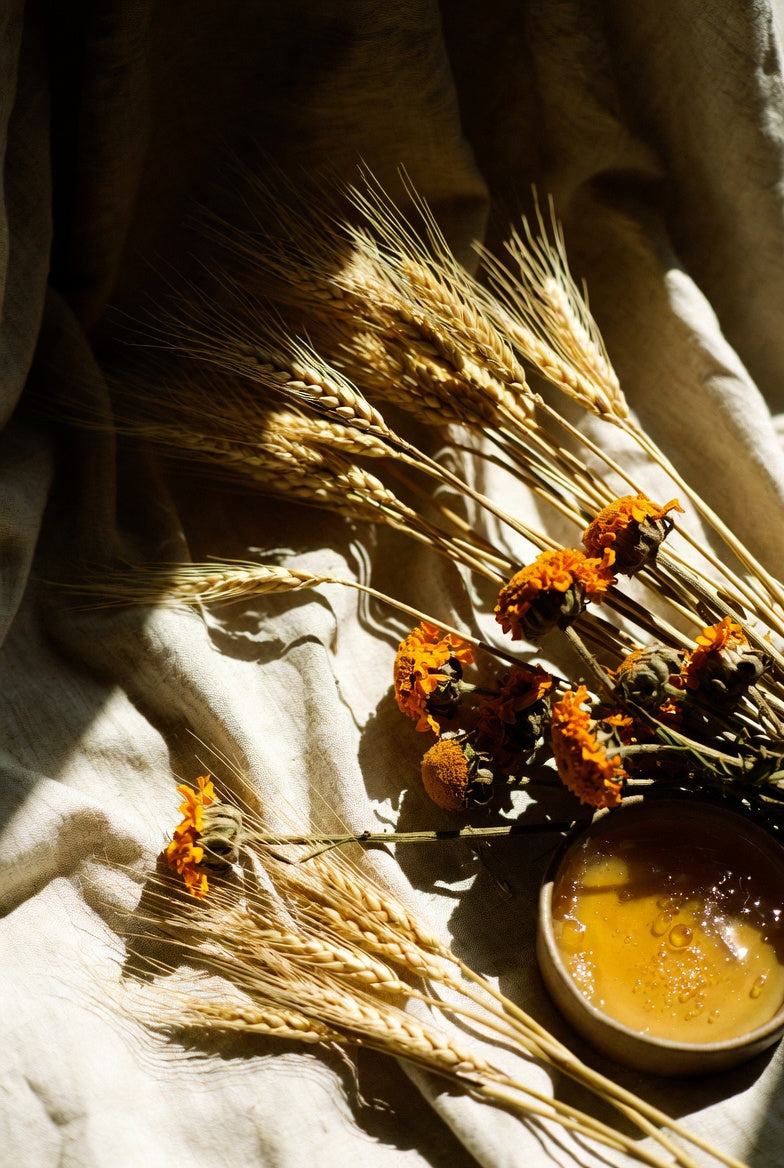 Sun-warmed golden wheat and dried marigold arranged on rumpled oat linen beside a small vessel of honey, late-afternoon light