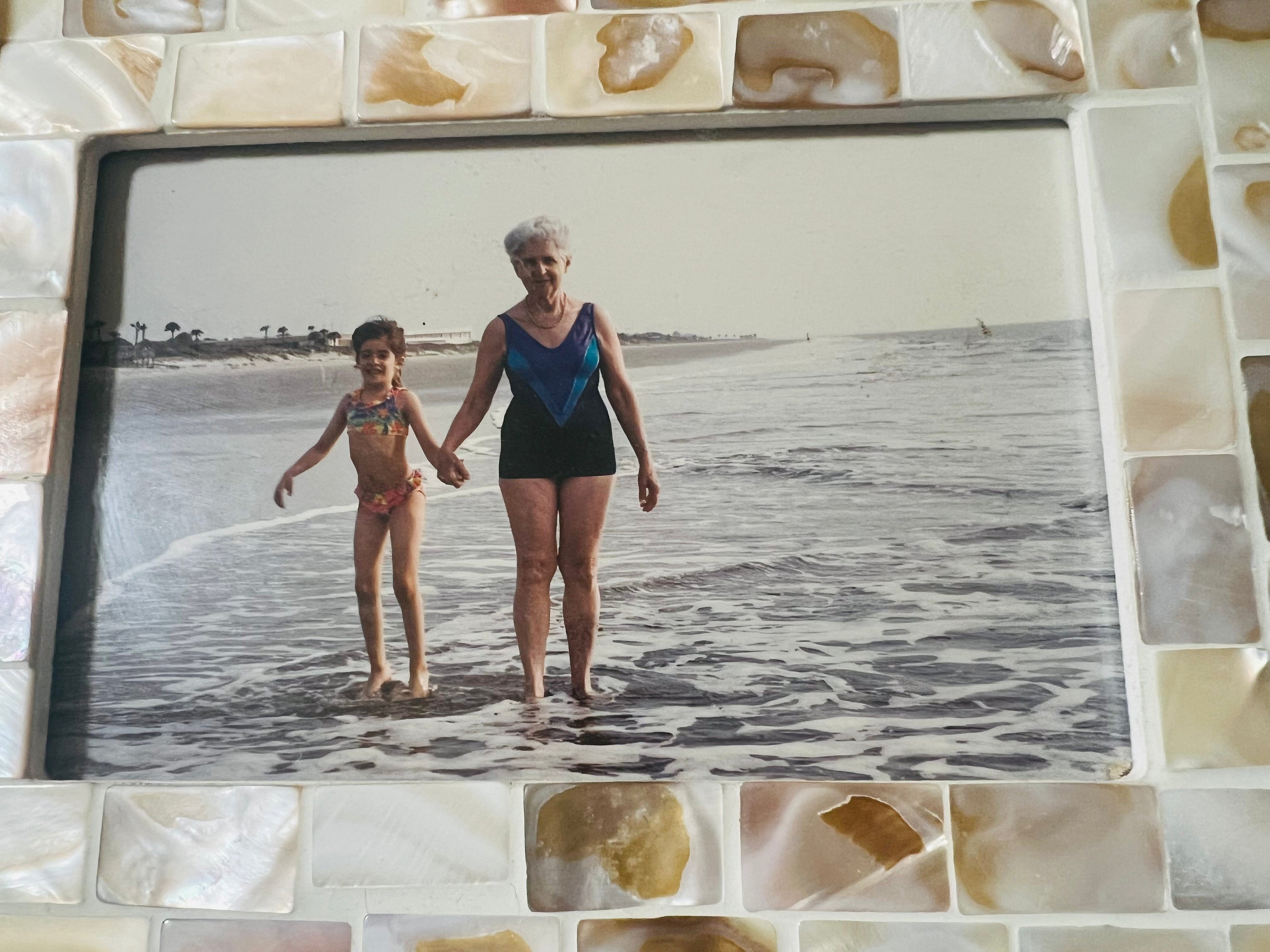 Young Stephanie Lyra holding her grandmother's hand at the ocean's edge, both in swimsuits with the shoreline behind them.
